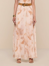A person is wearing the Torin Pleated Maxi Skirt—a sleeveless, light peach and beige poly chiffon tie-dye pleated maxi skirt with a brown belt—paired with a bracelet and beige open-toe heels, standing against a plain white background.
