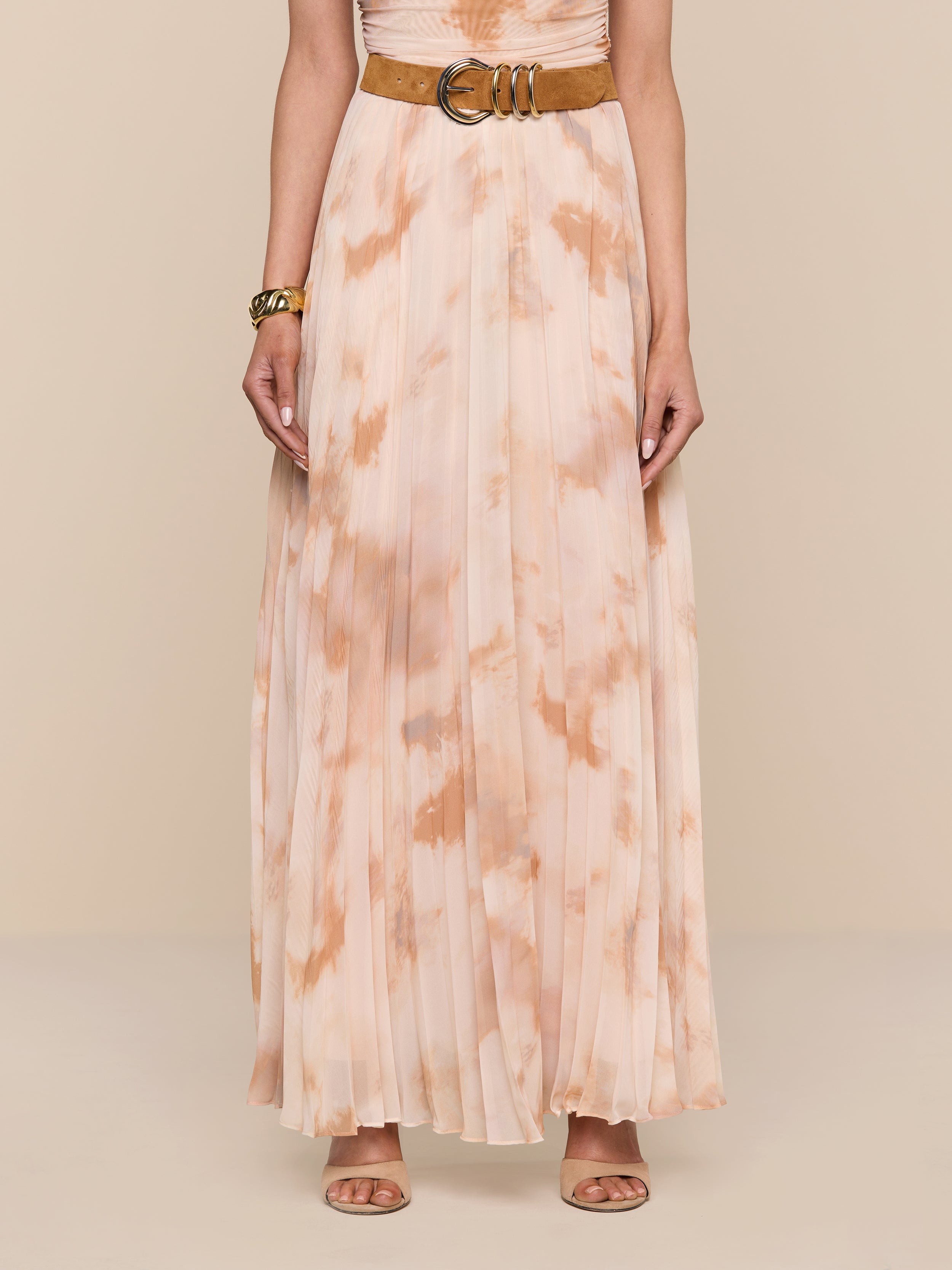 A person is wearing the Torin Pleated Maxi Skirt—a sleeveless, light peach and beige poly chiffon tie-dye pleated maxi skirt with a brown belt—paired with a bracelet and beige open-toe heels, standing against a plain white background.