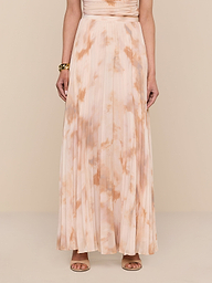 A person wearing the Torin Pleated Maxi Skirt, a floor-length, peach and beige poly chiffon tie-dye skirt with pleats, paired with tan heeled sandals stands against a white background. Only the person’s body from shoulders down is visible.