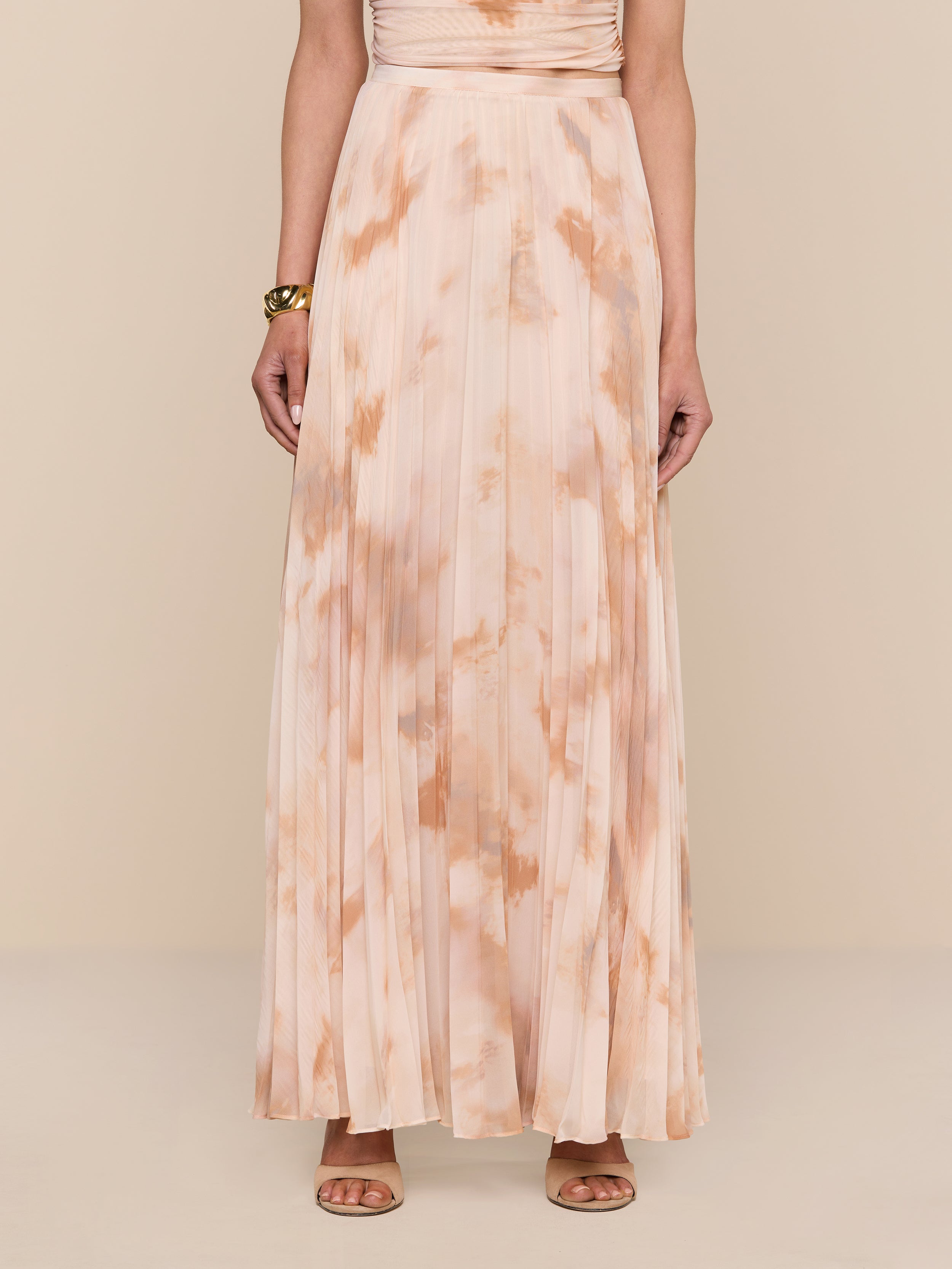 A person wearing the Torin Pleated Maxi Skirt, a floor-length, peach and beige poly chiffon tie-dye skirt with pleats, paired with tan heeled sandals stands against a white background. Only the person’s body from shoulders down is visible.