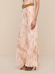 Wearing the Torin Pleated Maxi Skirt in a floor-length, light pink and beige tie-dye poly chiffon, styled with a sleeveless top, a brown belt, and brown heeled sandals, a person stands against a plain white background.