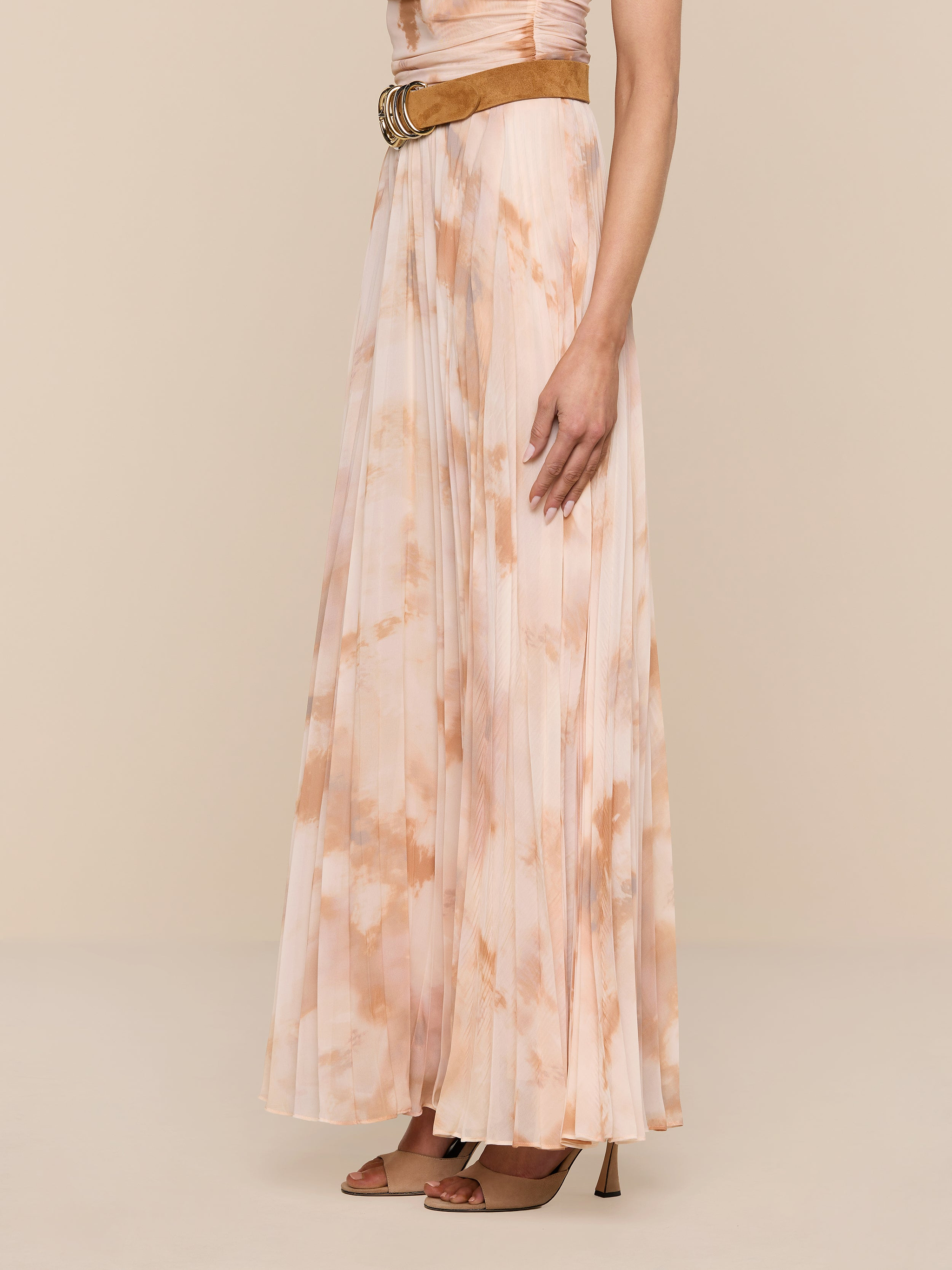 Wearing the Torin Pleated Maxi Skirt in a floor-length, light pink and beige tie-dye poly chiffon, styled with a sleeveless top, a brown belt, and brown heeled sandals, a person stands against a plain white background.