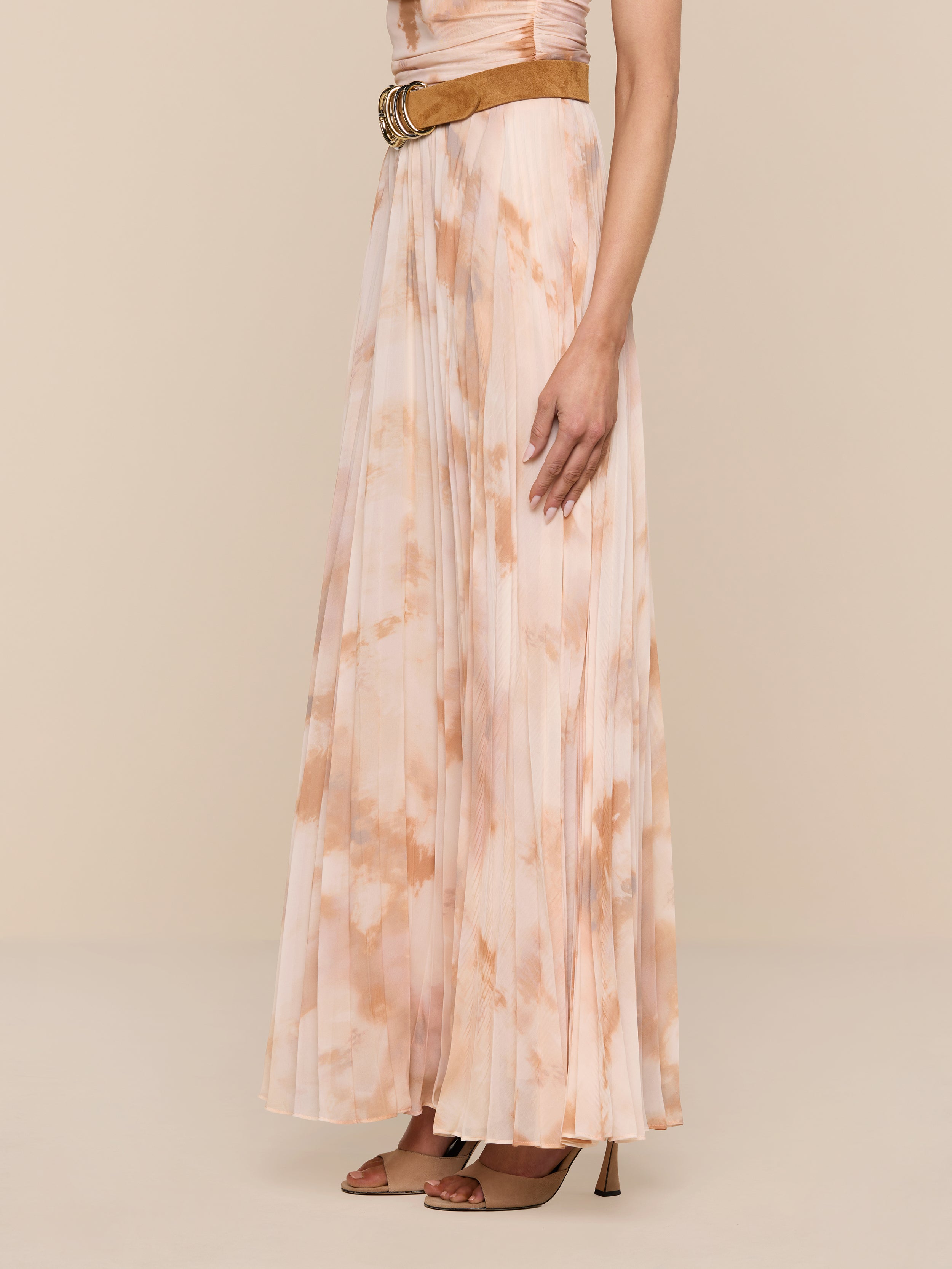 Wearing the Torin Pleated Maxi Skirt in a floor-length, light pink and beige tie-dye poly chiffon, styled with a sleeveless top, a brown belt, and brown heeled sandals, a person stands against a plain white background.