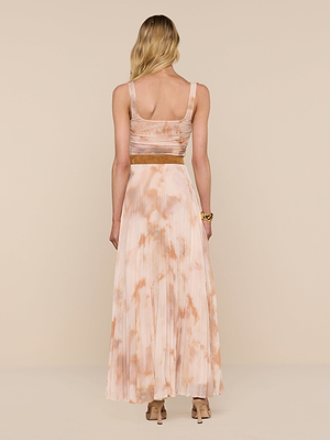 A woman with long blonde hair stands facing away, wearing the Torin Pleated Maxi Skirt in a pink and tan tie-dye pattern, paired with beige heels and a sleeveless top, against a plain light beige background.