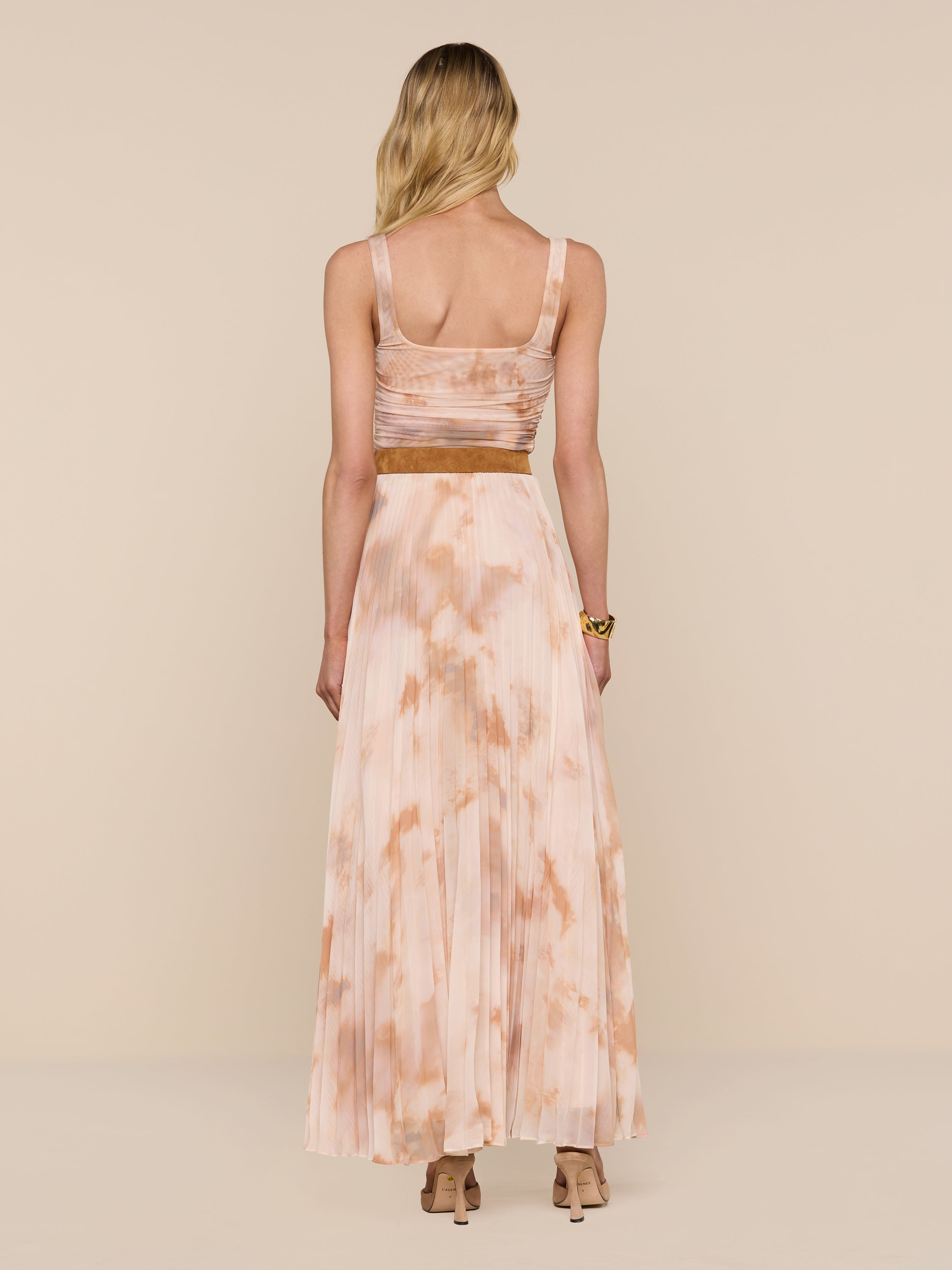 A woman with long blonde hair, wearing the Torin Pleated Maxi Skirt in a sleeveless light pink and beige tie-dye style, paired with heeled sandals, stands facing away against a plain white background.