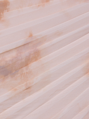 A close-up of the Torin Pleated Maxi Skirt in light pink poly chiffon, showcasing diagonal pleats and subtle brownish stains that create a textured, slightly uneven appearance.