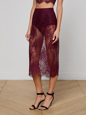 A person wears the L'AGENCE Makena Lace Pencil Skirt, a sheer floral lace burgundy midi with matching high-waisted shorts, paired with black ankle-strap heels, while standing on a wooden floor.