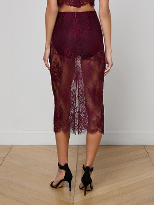 A woman stands on a wooden floor wearing the L'AGENCE Makena Lace Pencil Skirt in maroon, featuring sheer floral lace from waist to mid-calf that reveals matching undergarments. She is seen from the back in black high-heeled sandals.