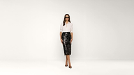 Woman wearing sunglasses, a white shirt, and a black sequin skirt stands against a plain white background, facing forward with a neutral expression.
