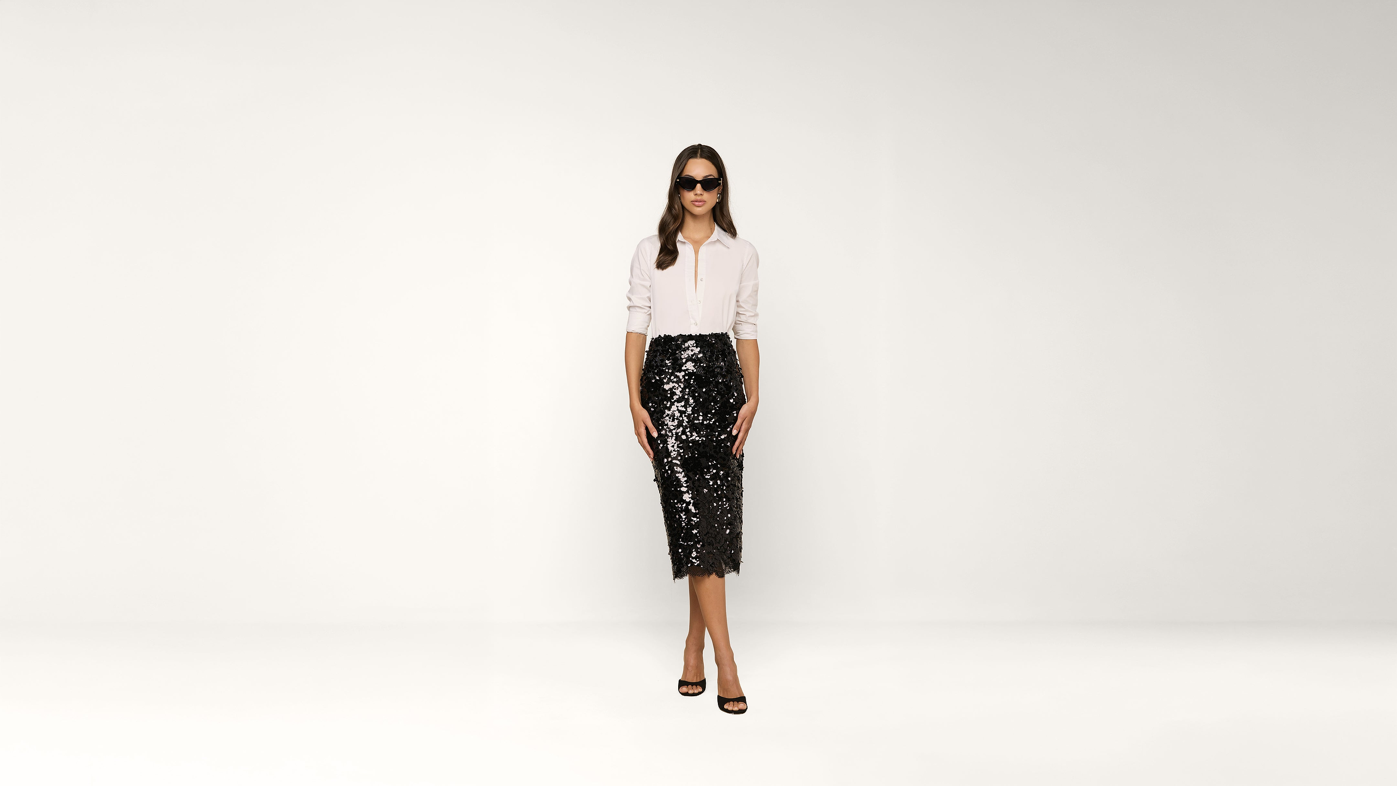 Woman wearing sunglasses, a white shirt, and a black sequin skirt stands against a plain white background, facing forward with a neutral expression.