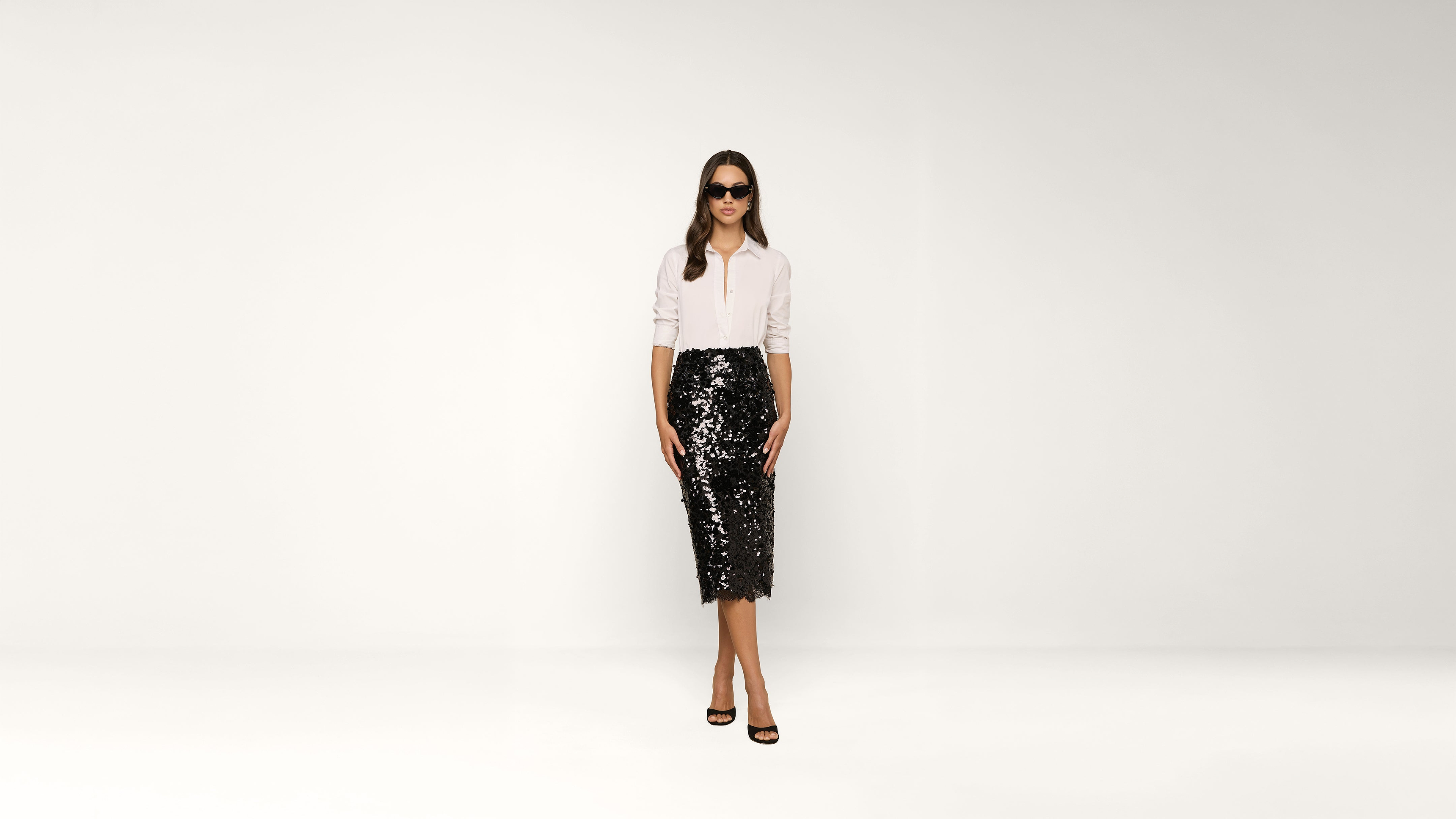 Woman wearing sunglasses, a white shirt, and a black sequin skirt stands against a plain white background, facing forward with a neutral expression.