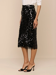 Eine Frau steht vor einem beigen Hintergrund in einem weißen, langärmeligen Hemd, schwarzen, offenen Schuhen und dem Makena Pailletten-Bleistiftrock - ein eleganter Look, perfekt für formelle Anlässe oder einen stilvollen Abend.