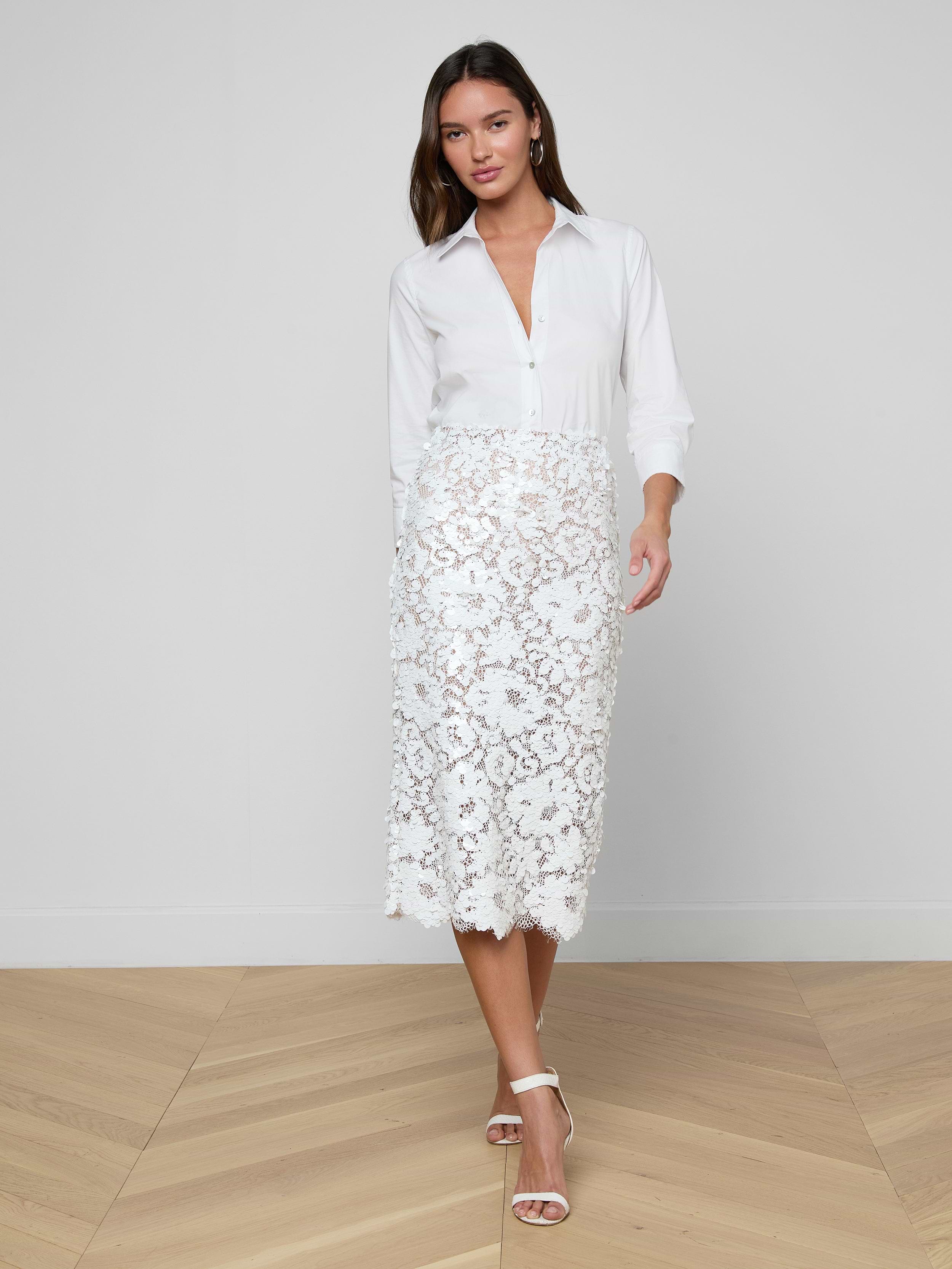 A woman wearing a white button-up blouse and the Makena Sequin Pencil Skirt stands against a plain light background, posing with one foot forward. She completes the look with white heeled sandals, embracing fresh spring fashion.