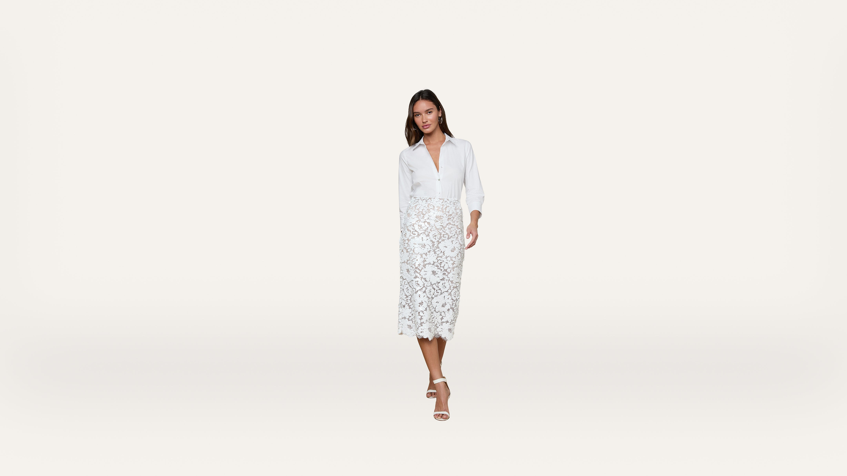 A woman in a white blouse and white lace midi skirt stands against a plain white background, wearing white heeled sandals.