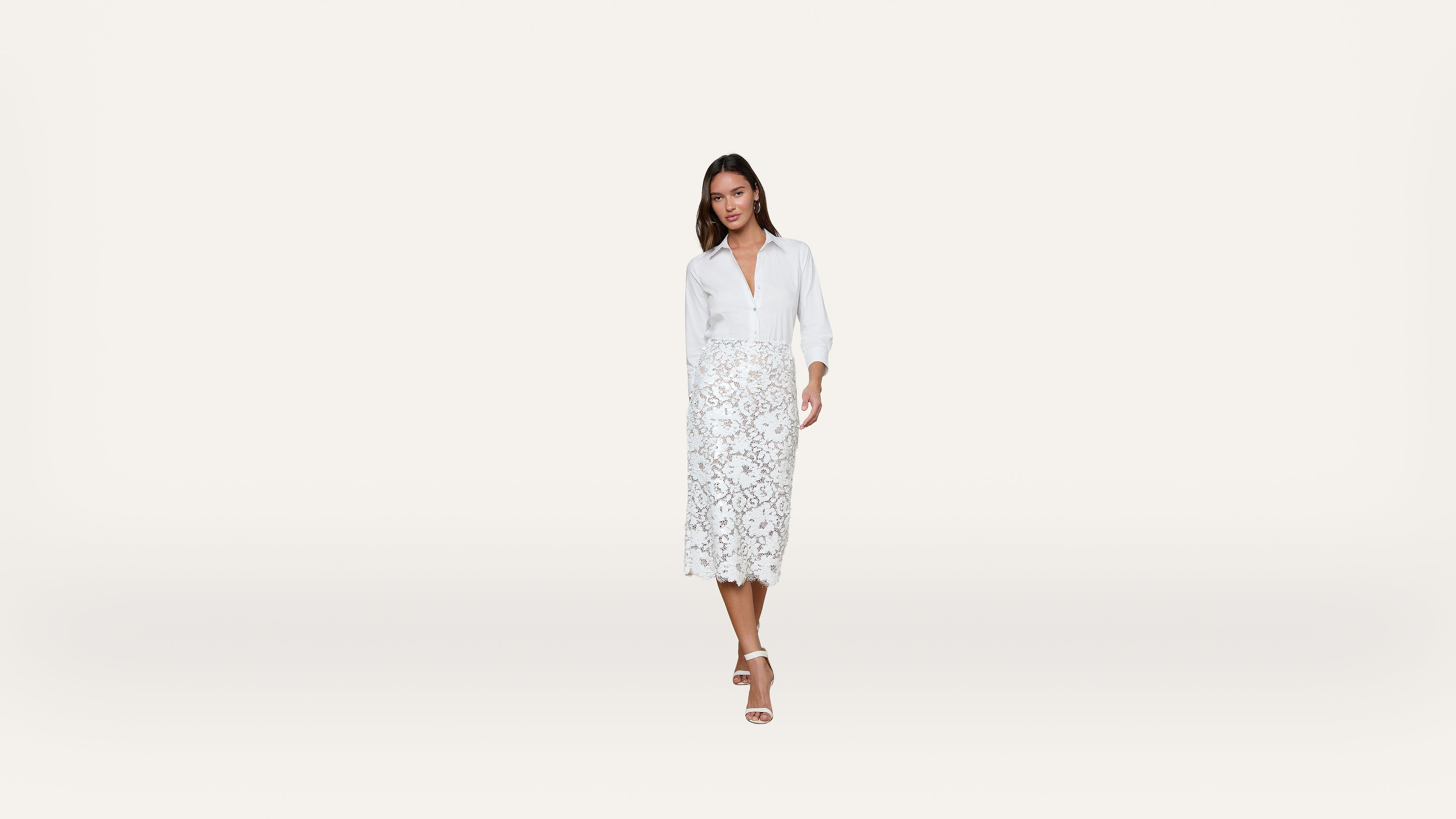 A woman in a white blouse and white lace midi skirt stands against a plain white background, wearing white heeled sandals.