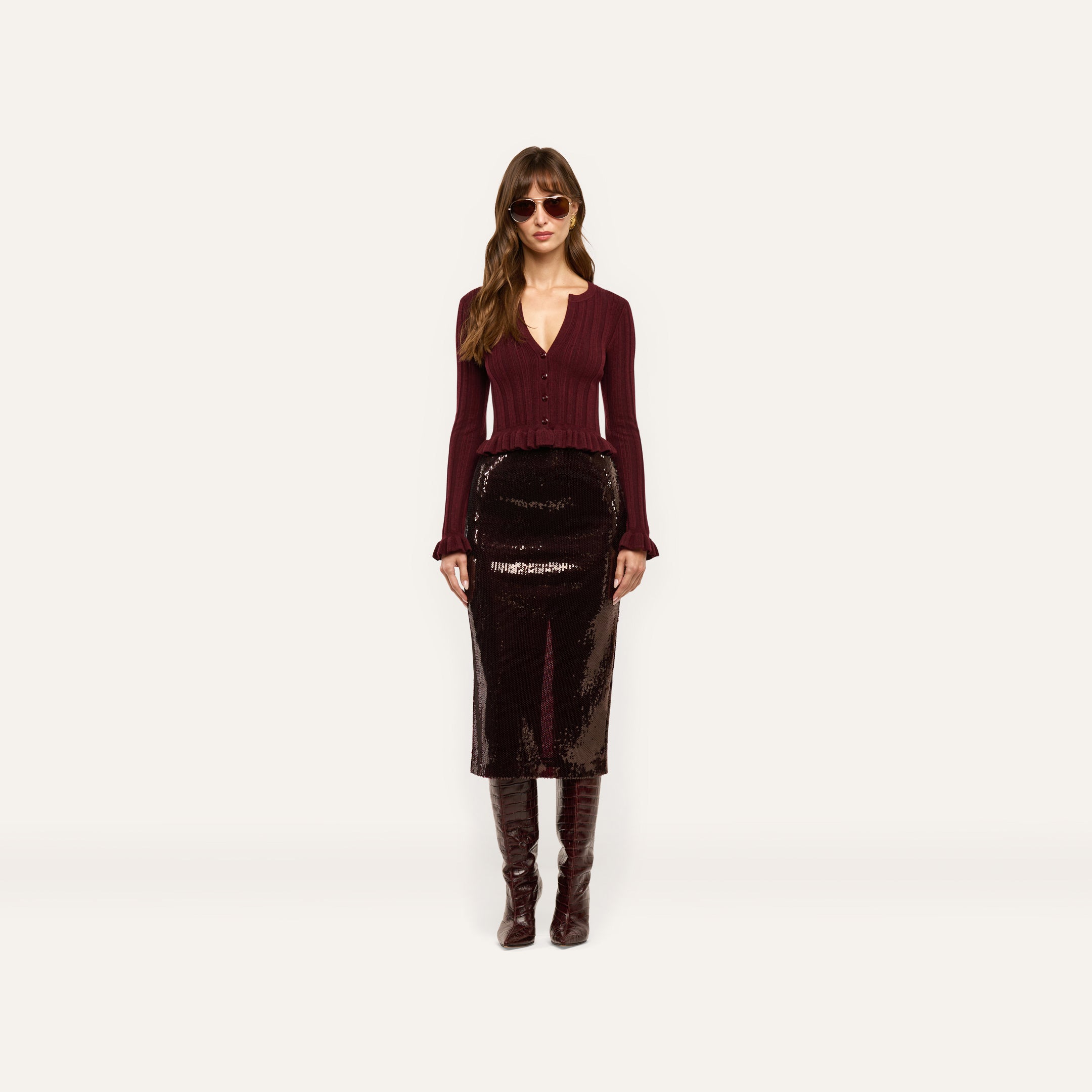 A model wears a long, espresso brown Makena Sequin Pencil Skirt, a burgundy button-down top with ruffled cuffs, and knee-high boots.