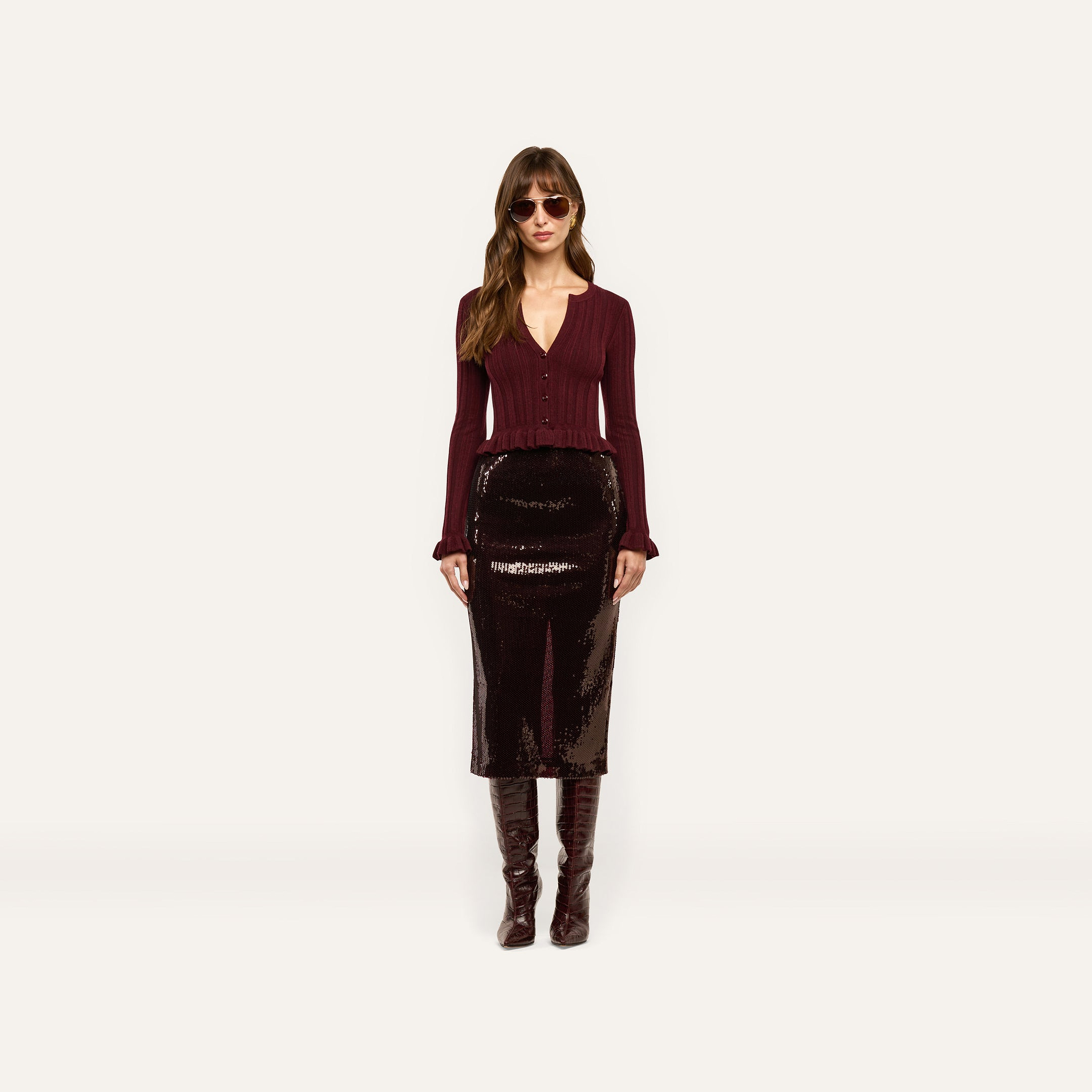 A model wears a long, espresso brown Makena Sequin Pencil Skirt, a burgundy button-down top with ruffled cuffs, and knee-high boots.