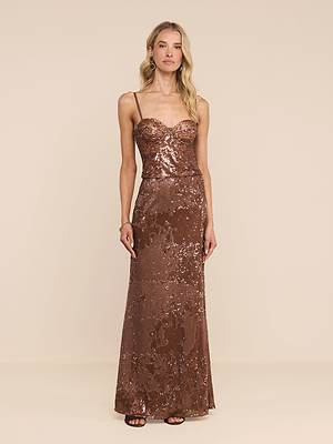 A woman stands against a plain beige background, looking forward with a neutral expression while wearing the Zeta Sequin Maxi Skirt, a fitted, floor-length bronze skirt adorned with sequins.