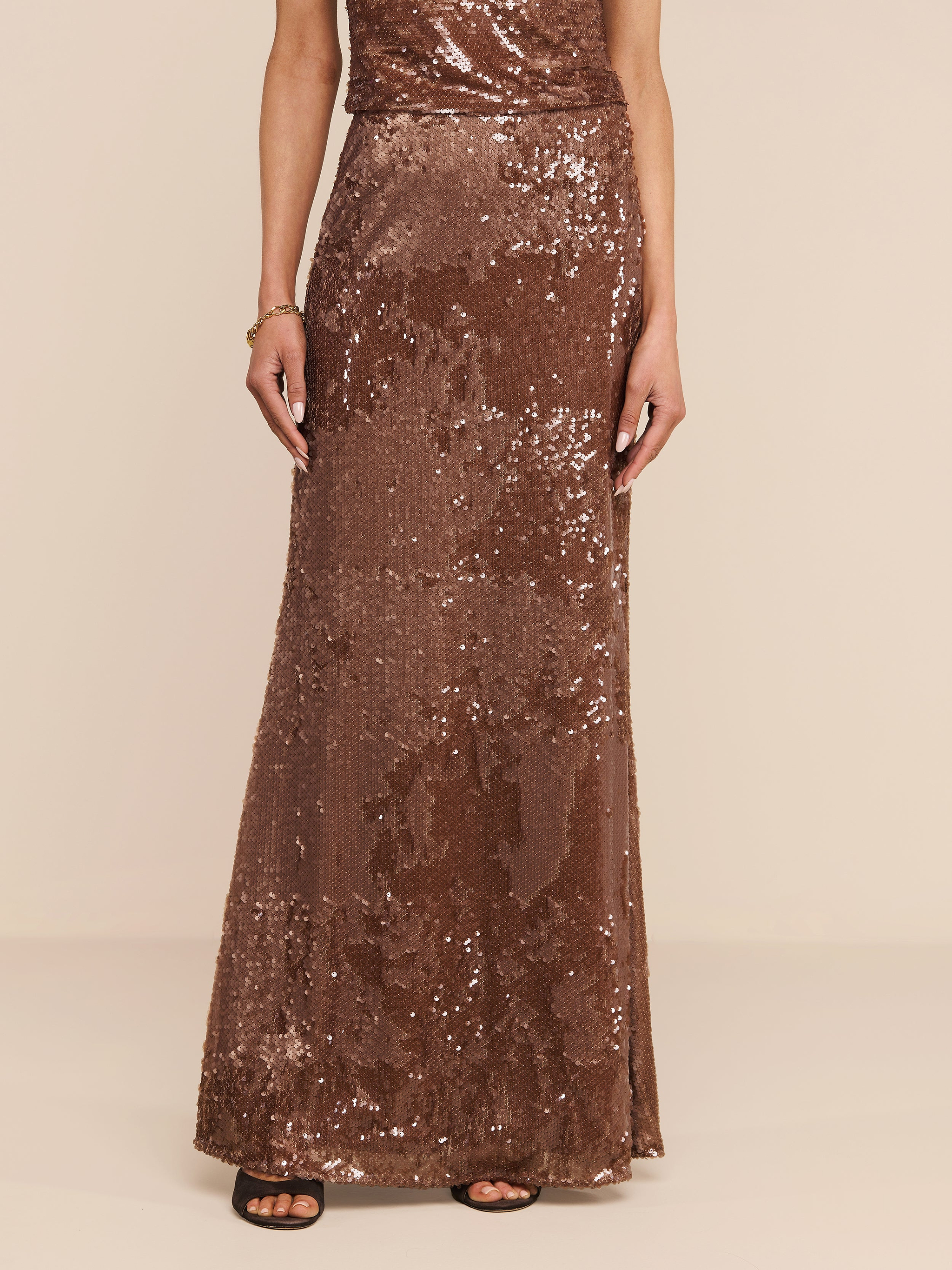A person wearing the Zeta Sequin Maxi Skirt, a floor-length, sleeveless brown sequin skirt by L’AGENCE, paired with black open-toed heels stands against a plain white background. The photo is cropped at the shoulders.