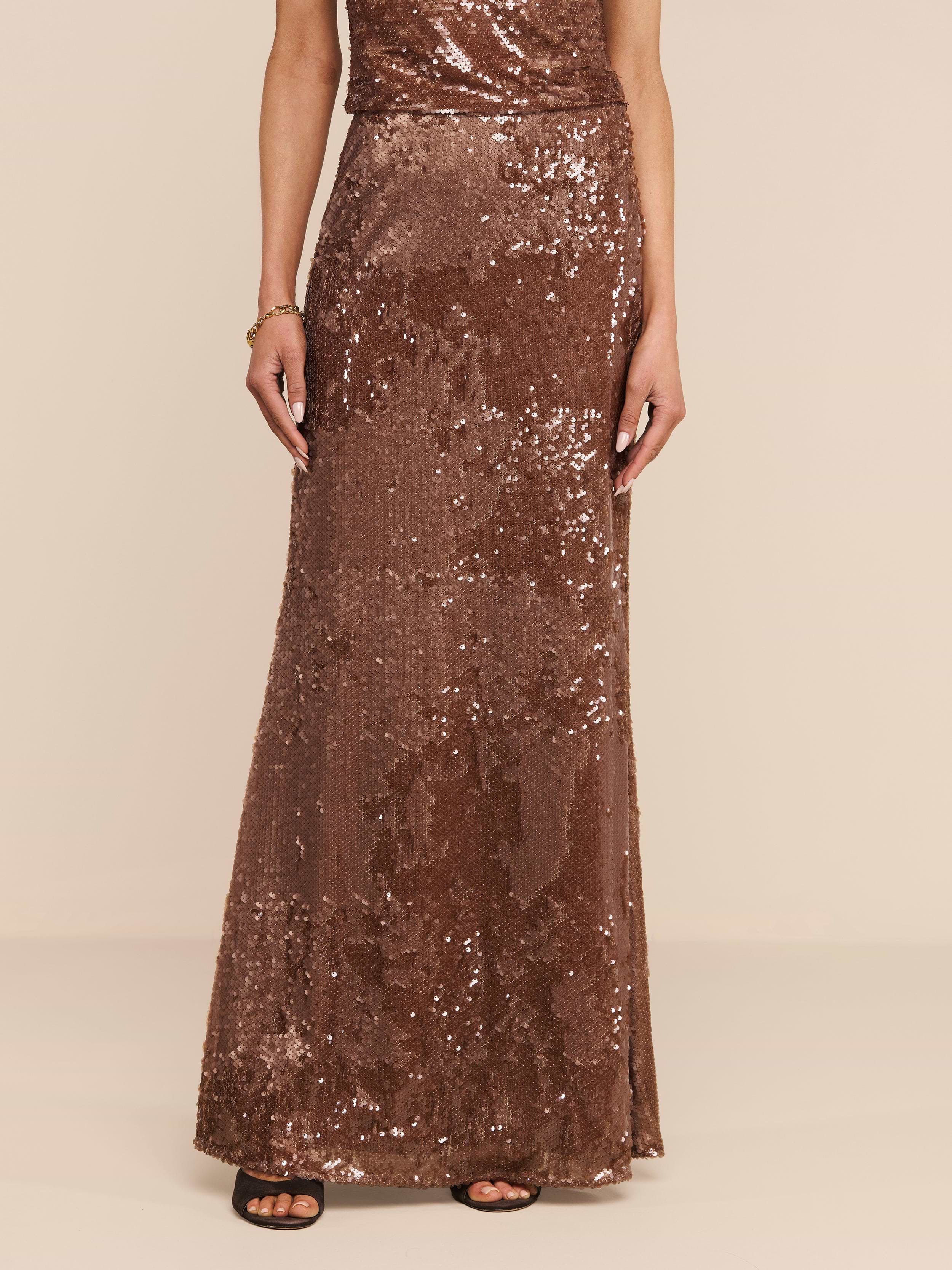 A person wearing the Zeta Sequin Maxi Skirt, a floor-length, sleeveless brown sequin skirt by L’AGENCE, paired with black open-toed heels stands against a plain white background. The photo is cropped at the shoulders.