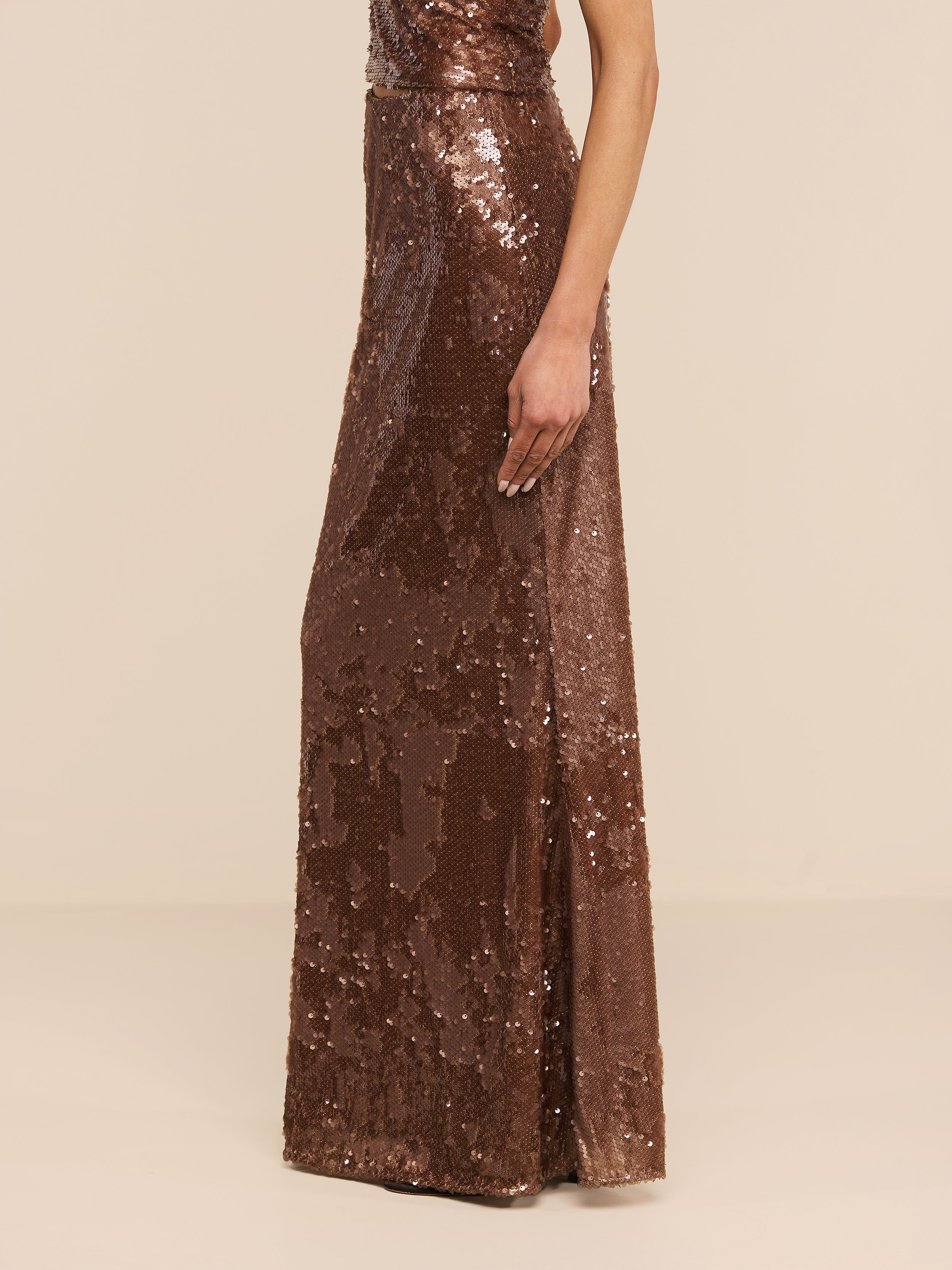 A person wearing the Zeta Sequin Maxi Skirt, a sleeveless, floor-length brown sequin skirt, stands against a plain white background with their head out of frame.