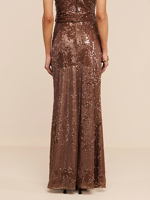 A person is shown from the back, standing against a plain beige background, wearing the Zeta Sequin Maxi Skirt by L’AGENCE in brown.