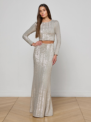 A woman with long straight hair stands on a wooden floor against a plain light background, wearing a silver long-sleeve sequin crop top and the L’AGENCE Zeta Sequin Maxi Skirt, her left hand resting on her hip.