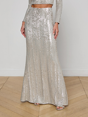 A person wears the L’AGENCE Zeta Sequin Maxi Skirt with a matching long-sleeve crop top, standing on a light wood floor against a plain white wall.