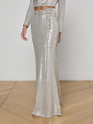 A person wearing the L'AGENCE Zeta Sequin Maxi Skirt with a matching long-sleeve crop top stands on a wooden floor against a plain white wall.
