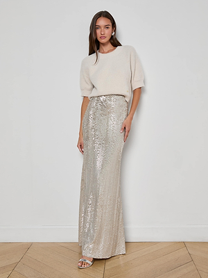 A woman with straight brown hair and a neutral expression stands against a white wall, wearing a cream short-sleeve sweater, L'AGENCE Zeta Sequin Maxi Skirt in silver, and silver sandals on a light wood floor.