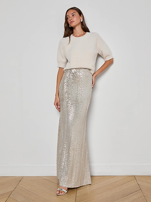 A woman with long hair and minimal accessories stands against a white wall, wearing a fuzzy cream short-sleeved sweater, silver sandals, and the L'AGENCE Zeta Sequin Maxi Skirt. Her expression is neutral.
