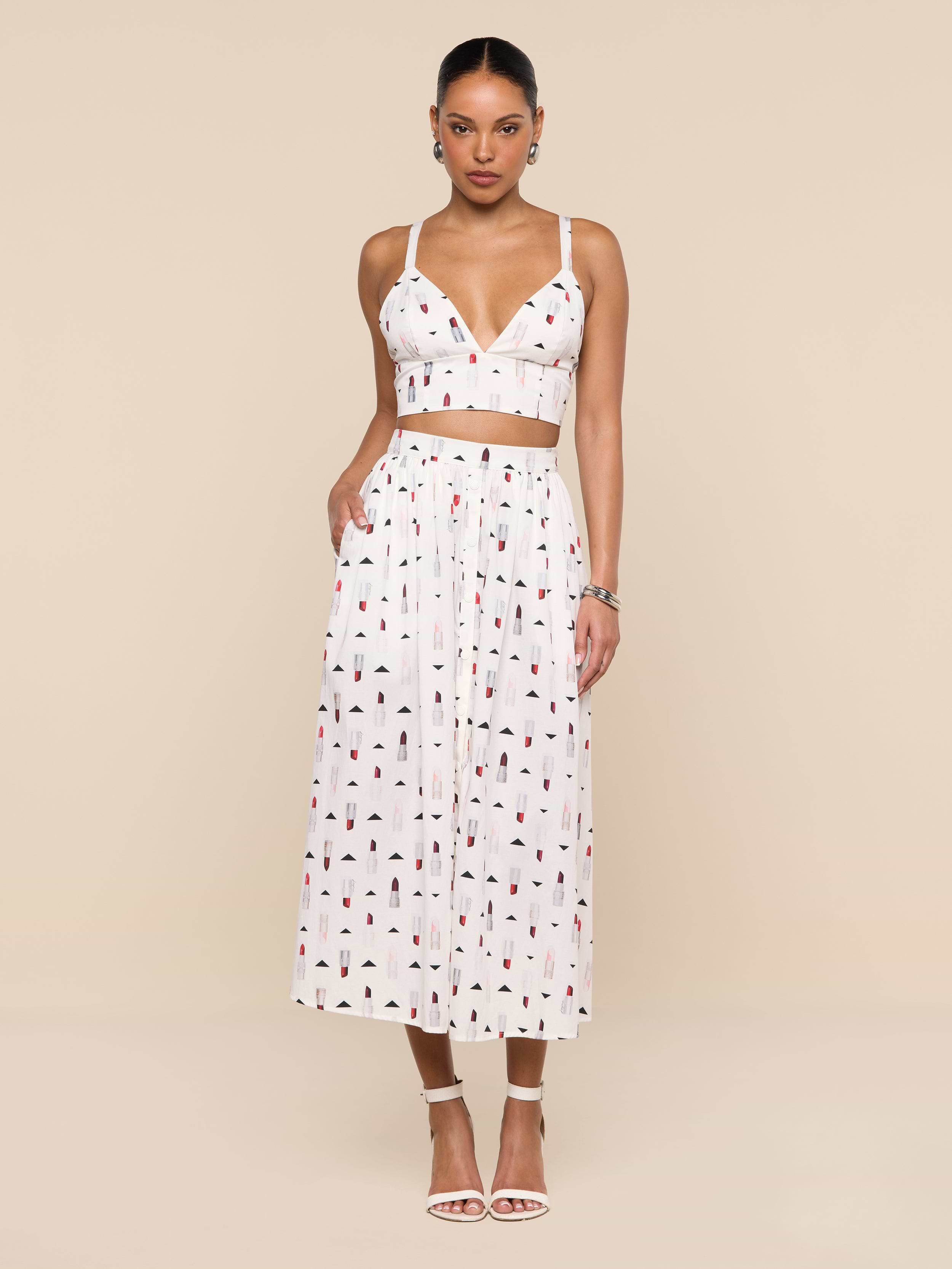 A woman stands against a white background wearing a white crop top and the Seina Poplin Midi Skirt—a high-waisted, ankle-length skirt featuring a small colorful print—paired with white high-heeled sandals.