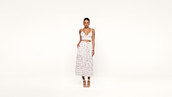 A woman stands against a plain white background, wearing a white, sleeveless, v-neck crop top and a matching high-waisted, mid-length skirt with a small, colorful print. She is also wearing white heels.