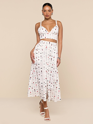 A woman wearing a white sleeveless crop top and the Seina Poplin Midi Skirt with a colorful print, paired with white strappy heels and hoop earrings, stands against a beige background.