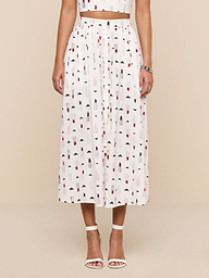 A woman stands against a plain white background wearing the Seina Poplin Midi Skirt, a high-waisted white skirt with a colorful geometric print, paired with a matching crop top, white ankle-strap heels, and a silver bracelet.