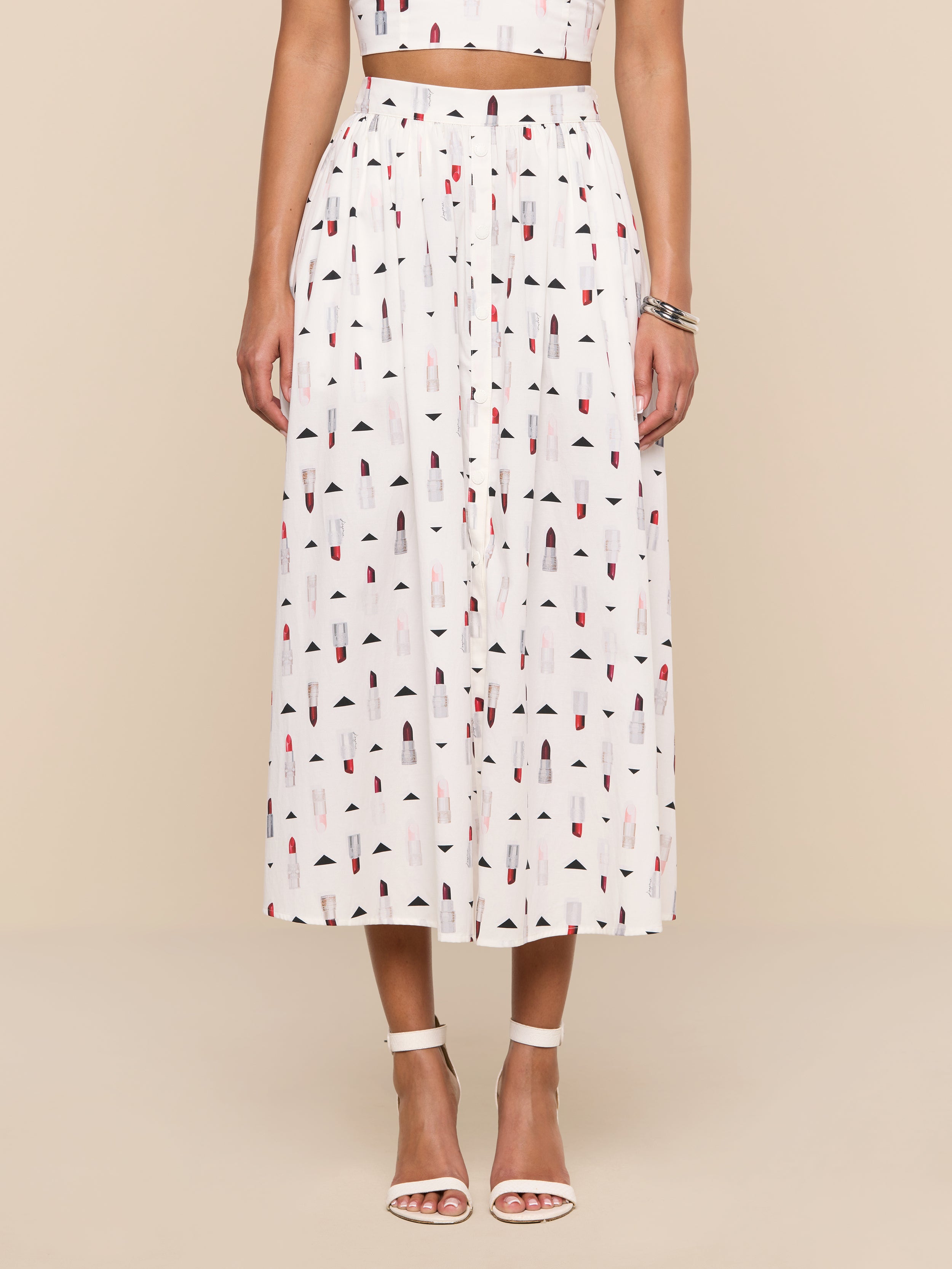 A woman stands against a plain white background wearing the Seina Poplin Midi Skirt, a high-waisted white skirt with a colorful geometric print, paired with a matching crop top, white ankle-strap heels, and a silver bracelet.