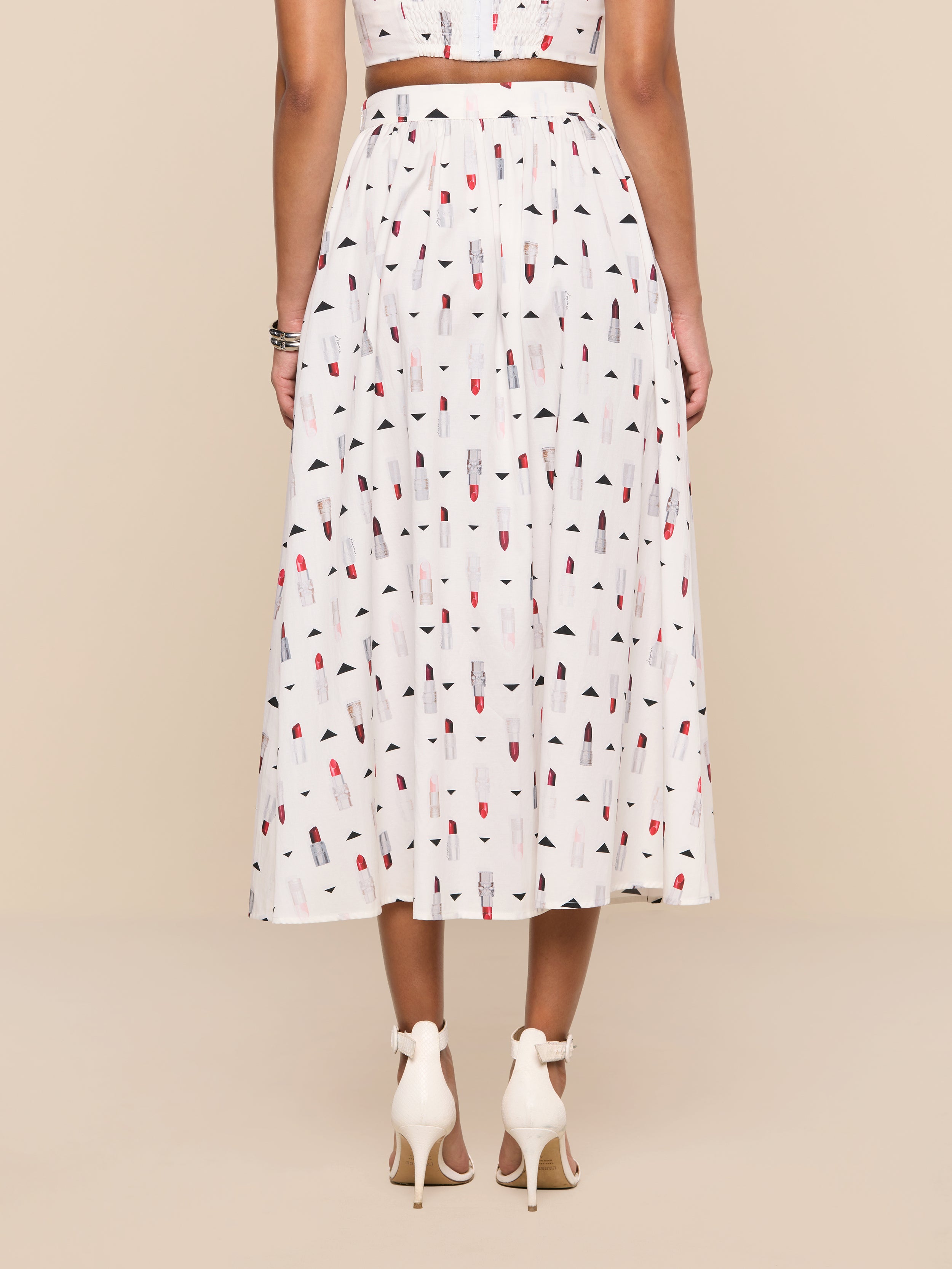 A woman stands facing away, wearing the Seina Poplin Midi Skirt—a white pleated skirt adorned with a colorful popsicle print—paired with white high-heeled sandals. The background is plain white.