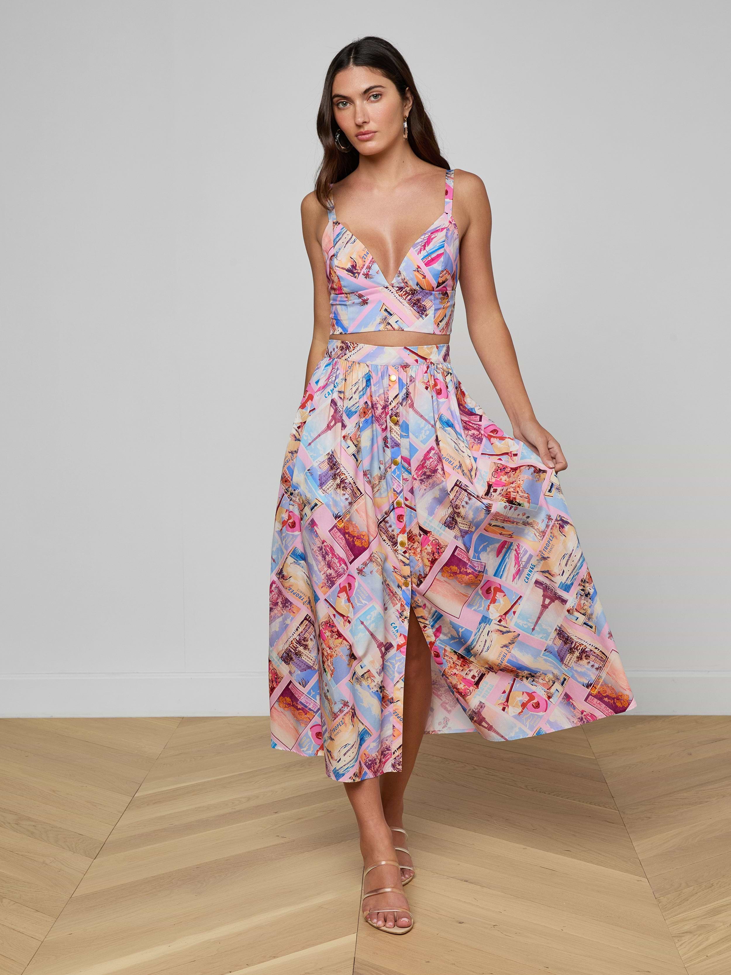 A woman stands indoors on wooden flooring against a plain light gray wall, wearing a colorful postcard print crop top and the Seina Poplin Midi Skirt. She is holding one side of her high-waisted, mid-length skirt.
