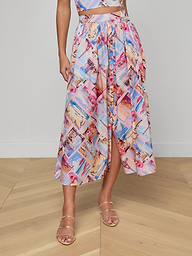 A person wears the Seina Poplin Midi Skirt, a colorful high-waisted piece with postcard prints, front button detail, and a slight slit, styled with strappy gold sandals.
