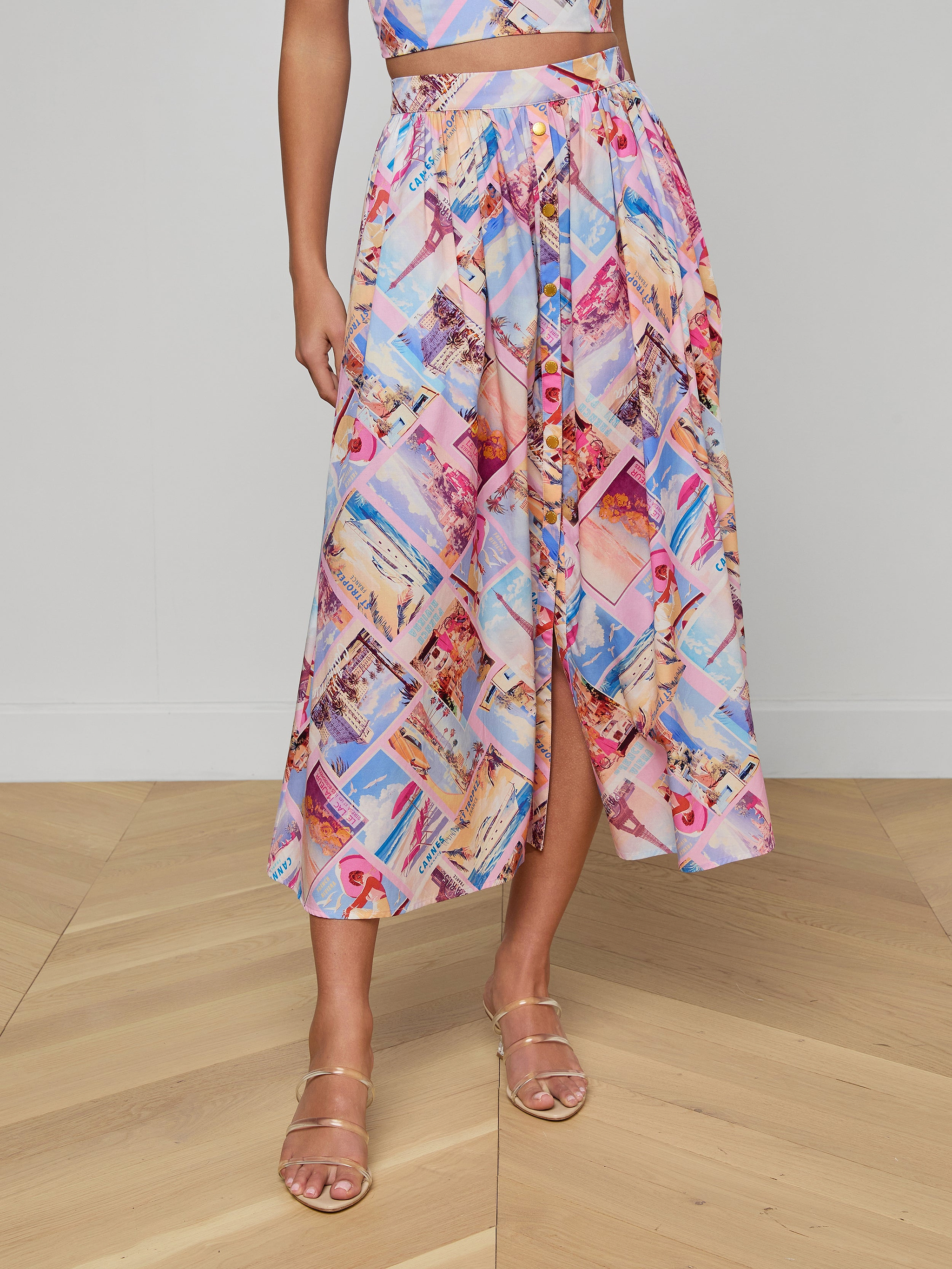 A person wears the Seina Poplin Midi Skirt, a colorful high-waisted piece with postcard prints, front button detail, and a slight slit, styled with strappy gold sandals.