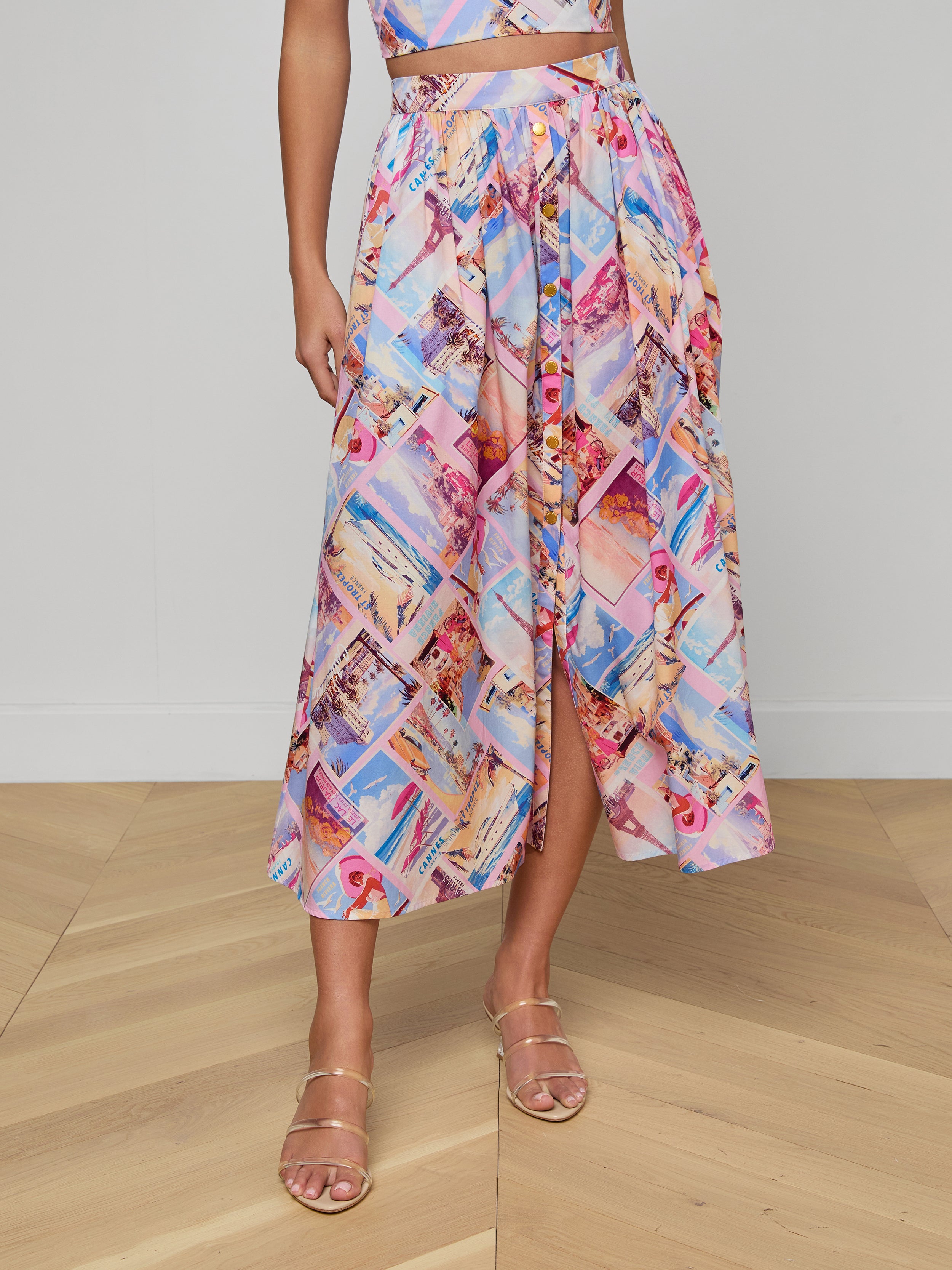 A person wears the Seina Poplin Midi Skirt, a colorful high-waisted piece with postcard prints, front button detail, and a slight slit, styled with strappy gold sandals.