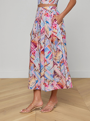 A person wearing the Seina Poplin Midi Skirt in a colorful postcard print, a matching crop top, and clear strappy sandals stands on a light wood floor against a plain white wall.
