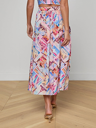 A woman stands indoors on a wooden floor, photographed from behind, wearing the Seina Poplin Midi Skirt with a colorful postcard print and nude open-heeled sandals.