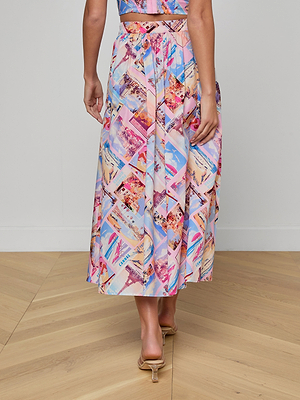 A woman stands indoors on a wooden floor, photographed from behind, wearing the Seina Poplin Midi Skirt with a colorful postcard print and nude open-heeled sandals.