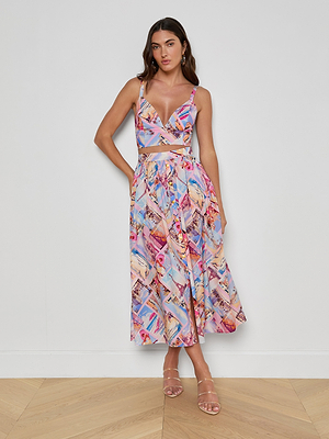 A woman with long brown hair stands against a white wall, wearing the Seina Poplin Midi Skirt featuring a colorful postcard print and a flowy silhouette, paired with strappy nude heeled sandals.