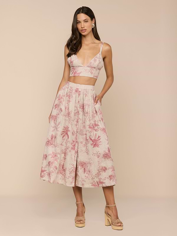 A woman poses in a sleeveless, pink and white jungle toile print crop top with a deep V-neck, paired with the Seina Linen Midi Skirt. She completes the look with nude platform sandals against a beige background.