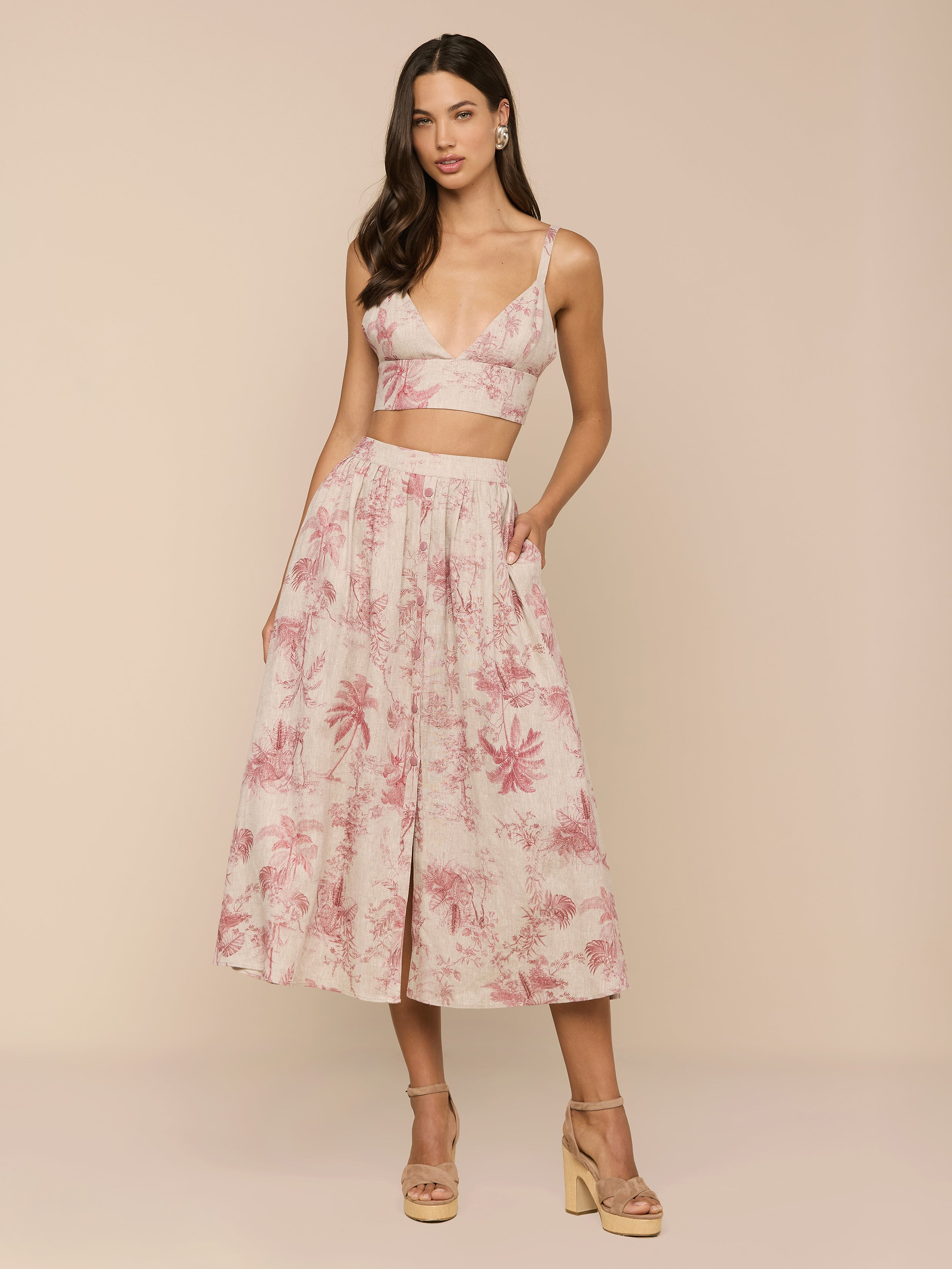 A woman poses in a sleeveless, pink and white jungle toile print crop top with a deep V-neck, paired with the Seina Linen Midi Skirt. She completes the look with nude platform sandals against a beige background.