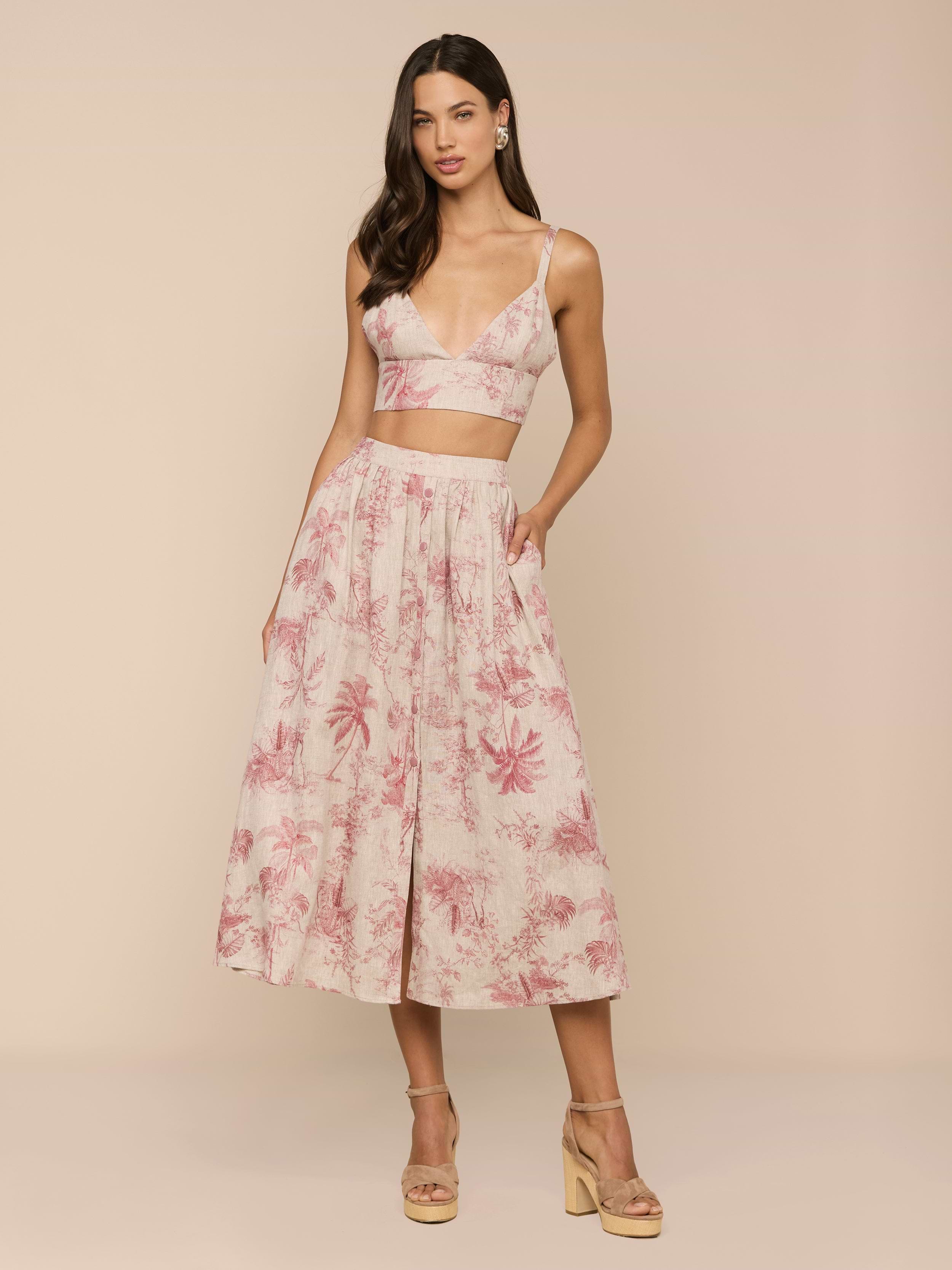 A woman poses in a sleeveless, pink and white jungle toile print crop top with a deep V-neck, paired with the Seina Linen Midi Skirt. She completes the look with nude platform sandals against a beige background.