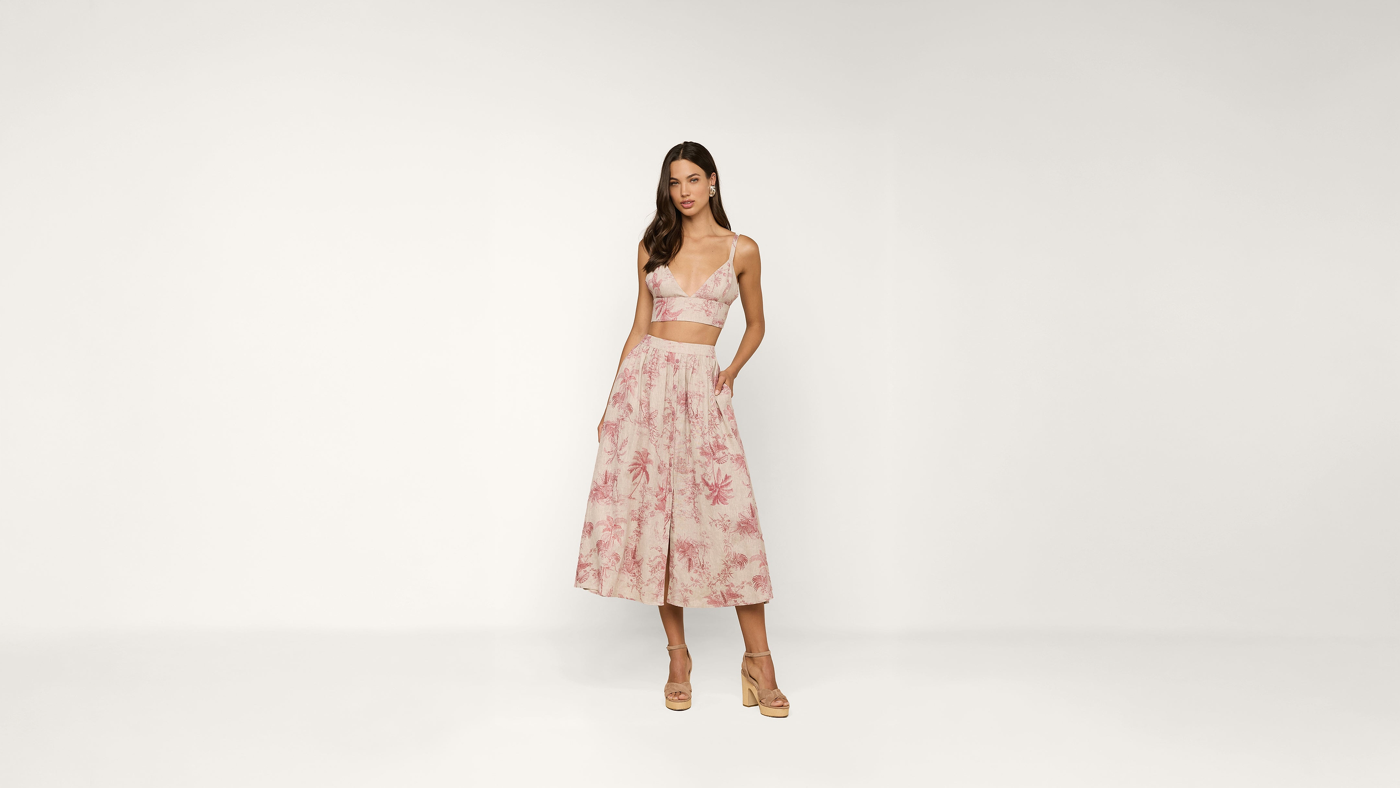 Woman wearing a light pink crop top and a matching floral midi skirt with pockets, paired with beige high-heeled sandals, standing against a plain white background.