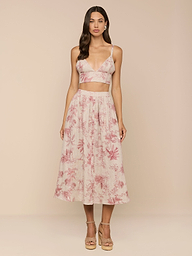 A woman stands against a beige background wearing the Seina Linen Midi Skirt, a high-waisted, flowy skirt with a pink floral pattern, paired with a sleeveless light crop top and beige platform heels.